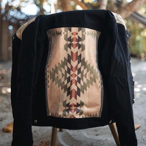 Pode incluir: Uma jaqueta de ganga preta com um painel traseiro estampado geométrico em tons de marrom, verde e bege.