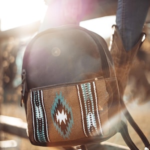 May include: A dark brown leather backpack featuring a front pocket with a woven textile panel. The panel displays a geometric pattern in shades of tan, turquoise, white, and black. The backpack includes a top zipper closure and adjustable straps.
