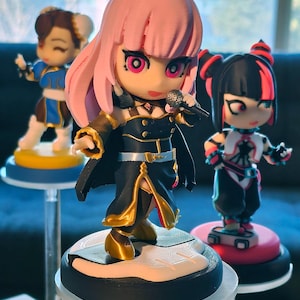 May include: Three anime-style figurines on clear stands. The central figure has pink hair, a black outfit with gold accents, and a microphone. Other figures include a blue and white outfit and a black and red outfit. The figurines are on round bases.