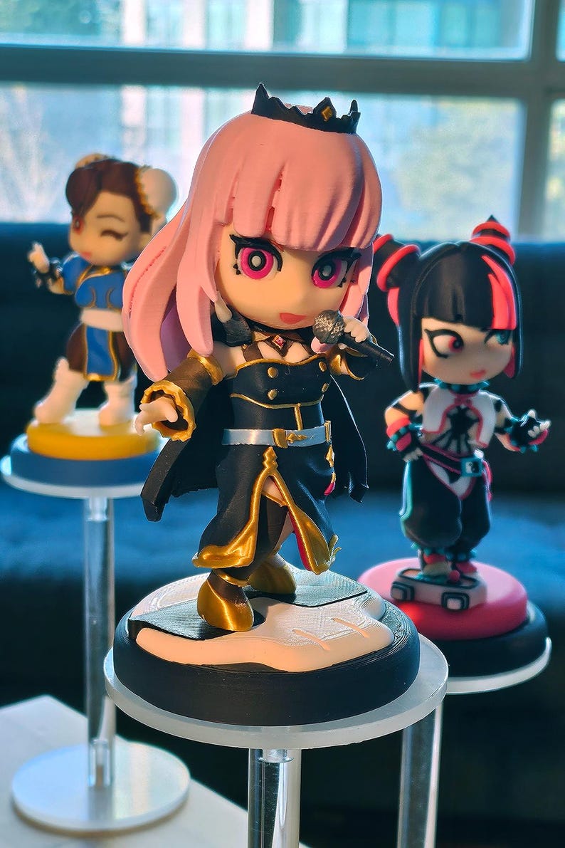 May include: Three anime-style figurines on clear stands. The central figure has pink hair, a black outfit with gold accents, and a microphone. Other figures include a blue and white outfit and a black and red outfit. The figurines are on round bases.