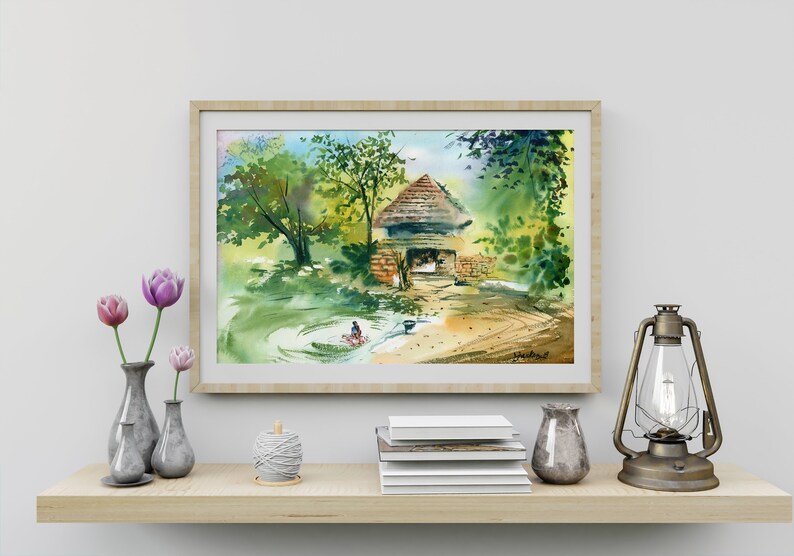 May include: A watercolor painting of a small, rustic house with a thatched roof, surrounded by lush green trees and a stream. The painting is framed in a light wood frame and is displayed on a shelf with other decorative items.