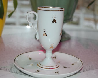May include: A white teacup and saucer set with gold trim and insect designs. The teacup has a unique handle and sits on a matching saucer. The set is likely porcelain and is a vintage or antique item.