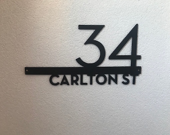 Puede incluir: Letrero de dirección de metal negro con el número 34 y el nombre de la calle "CARLTON ST". El letrero está montado en una pared texturizada de color claro. Los números y las letras están en una fuente sans-serif moderna.