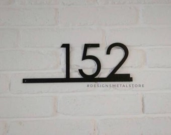 Puede incluir: Número de casa de metal negro 152 montado en una pared de ladrillos blancos. Los números están en una fuente moderna y conectados por una barra horizontal. El texto "#DESIGNSMETALSTORE" está debajo.