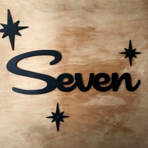 May include: Black metal sign spelling "Seven" in a cursive font, accompanied by four starburst-shaped accents. The sign is mounted on a light-colored wooden surface, creating a striking contrast. The design has a mid-century modern aesthetic.