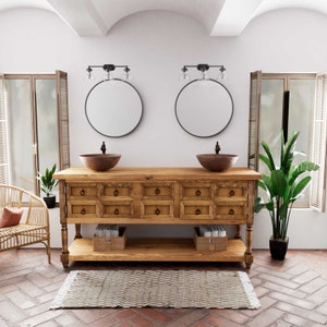 May include: A wooden bathroom vanity with two sinks, two round mirrors, and two light fixtures. The vanity has a shelf and drawers. The vanity is in a bathroom with a tiled floor and a plant.