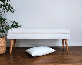 May include: White upholstered bench with wooden legs and a white throw pillow. The bench is sitting on a wooden floor.