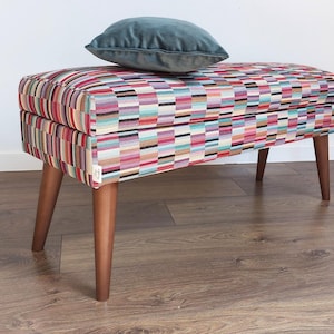 Könnte beinhalten: Eine farbenfrohe, gemusterte Aufbewahrungsbank mit Holzbeinen und einem türkisfarbenen Samtkissen darauf.