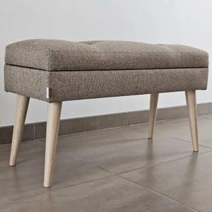 May include: A rectangular upholstered bench with a button-tufted top in a neutral brown fabric. The bench has four angled, light-colored wooden legs. The design is simple and modern, suitable for various interior styles.