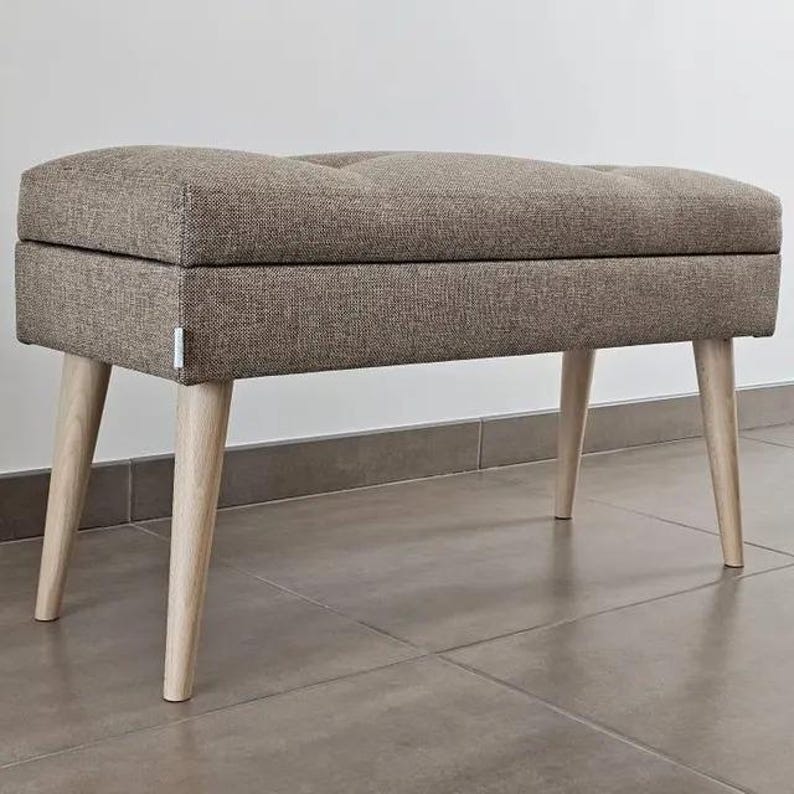 May include: A rectangular upholstered bench with a button-tufted top in a neutral brown fabric. The bench has four angled, light-colored wooden legs. The design is simple and modern, suitable for various interior styles.