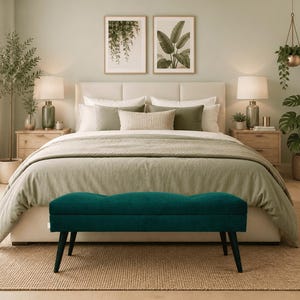 May include: A bedroom scene featuring a large bed with a neutral-toned comforter and pillows. A teal bench with black legs sits at the foot of the bed. Two framed botanical prints hang above the bed, and two wooden nightstands flank the bed, each with a lamp.