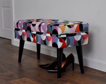 May include: A rectangular upholstered bench with a colorful geometric pattern in red, purple, blue, and orange. The bench has black legs and is paired with black high-heeled shoes. A stylish furniture piece.