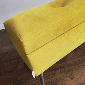 Può includere: Una panca rivestita in velluto giallo brillante con un design della seduta trapuntato e cuciture visibili lungo i bordi. La panca è sostenuta da sottili gambe in metallo scuro e presenta una piccola etichetta in tessuto bianco sul lato.