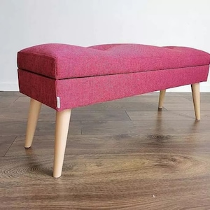Pode incluir: Um banco de armazenamento estofado rosa com pernas de madeira. O banco tem um topo estofado e uma forma retangular.