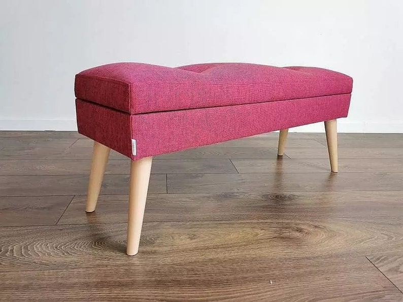 Pode incluir: Um banco de armazenamento estofado rosa com pernas de madeira. O banco tem um topo estofado e uma forma retangular.