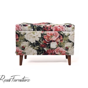 Op de afbeelding: Een opbergottoman met een bloemenpatroon en houten poten. De ottoman heeft een bloemenpatroon met roze, witte en groene bloemen.