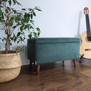 Może przedstawiać: Zielona welurowa ławka do przechowywania z drewnianymi nogami. Ławka znajduje się w pokoju z rośliną doniczkową i gitarą.