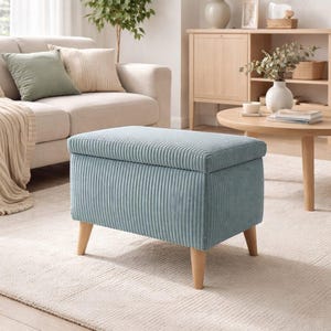 Puede incluir: Un otomán de pana azul claro con patas de madera. El banco de almacenamiento rectangular está en un salón, con un sofá y una alfombra beiges en el fondo. El otomán es un mueble elegante y funcional.