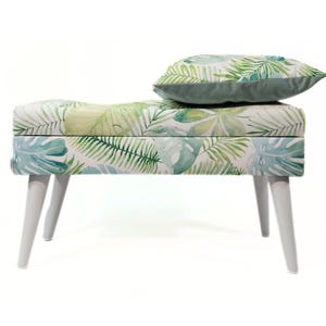 Op de afbeelding: Een witte bank met een botanische print met groene en blauwe bladeren. De bank heeft vier witte poten en een bijpassend kussen. Het kussen heeft een effen groene kleur. De bank is een stijlvol meubelstuk.