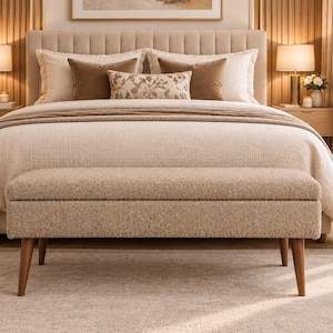 Op de afbeelding: Een rechthoekige opbergbank met een gestructureerde beige stoffen bekleding en vier taps toelopende houten poten. De bank staat aan het voeteneinde van een bed met een gecapitonneerd hoofdeinde, lichtgekleurd beddengoed en diverse sierkussens.