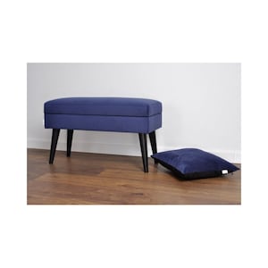 May include: A navy blue velvet storage bench with black wooden legs. The bench is sitting on a wooden floor and there is a matching navy blue pillow on the floor in front of it.