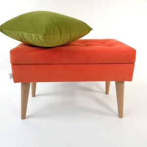 May include: A bright orange storage ottoman with a tufted top and wooden legs. A green pillow is placed on top of the ottoman.