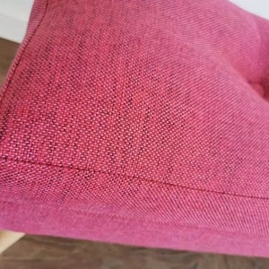 Pode incluir: Close-up de um pufe de tecido rosa com um bot&atilde;o de decora&ccedil;&atilde;o. O pufe tem um tecido texturizado e uma base de madeira.