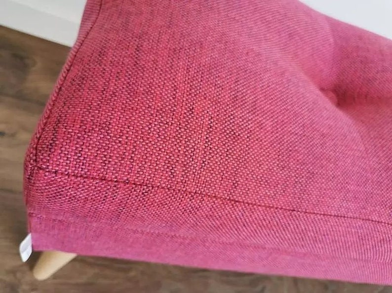 Pode incluir: Close-up de um pufe de tecido rosa com um bot&atilde;o de decora&ccedil;&atilde;o. O pufe tem um tecido texturizado e uma base de madeira.
