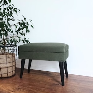 Può includere: Una panca di stoffa verde con gambe nere. La panca è appoggiata su un pavimento di legno davanti a un muro bianco.