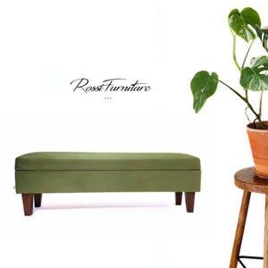 Pode incluir: Um banco estofado verde-oliva com pernas de madeira escura. O banco é retangular e tem uma textura macia e aveludada. A imagem também apresenta uma planta em vaso com folhas verdes grandes e um banco de madeira.