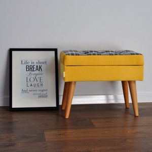 Op de afbeelding: Een gele opbergottoman met een zwart-wit geruite bovenkant. De ottomaan heeft vier houten poten en staat op een houten vloer. De tekst op de ingelijste print achter de ottomaan luidt: "Life is short Break Forgive Love Laugh And never regret anything that made you smile."