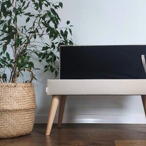 May include: A modern TV stand with a beige fabric-covered body and light wood legs. The stand supports a black screen. A woven basket with a plant is to the left. The stand is approximately 40 inches long.