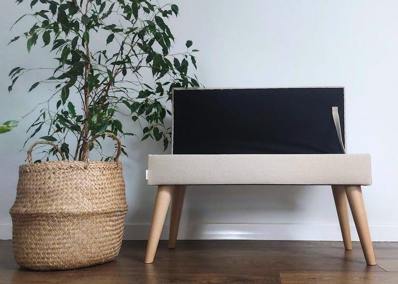 May include: A modern TV stand with a beige fabric-covered body and light wood legs. The stand supports a black screen. A woven basket with a plant is to the left. The stand is approximately 40 inches long.