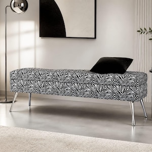 May include: A modern bench with a zebra print pattern in black and white. The bench has a rectangular shape with a tufted top and chrome legs. A black pillow rests on the bench. The bench is in a room with a floor lamp and a side table.