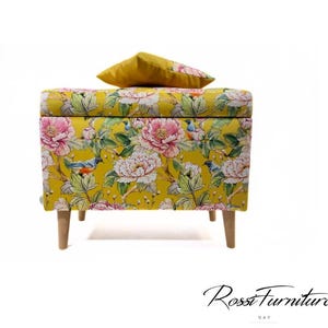 Könnte beinhalten: Eine gelbe Aufbewahrungskiste mit einem Blumendruck mit rosa Blumen und Vögeln. Die Kiste hat Holzbeine und ein gelbes Kissen oben drauf.