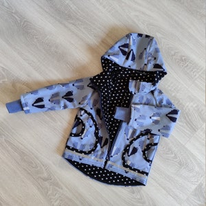 Può includere: Una giacca blu per bambini con stampa di tasso e fodera nera con stelle bianche. La giacca ha una chiusura a zip e un cappuccio.