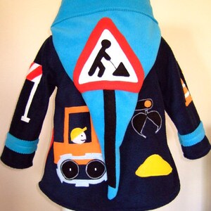 Könnte beinhalten: Eine blaue Fleecejacke mit Kapuze für Kinder mit Bau-Thema. Die Jacke zeigt ein gelbes Baufahrzeug, ein Straßenschild mit einem Arbeiter und einen Erdhaufen.
