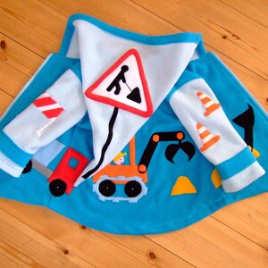 Könnte beinhalten: Eine blaue Fleecejacke mit Kapuze im Baustellen-Design. Die Jacke zeigt einen gelben Bagger, einen roten LKW, ein Straßenschild und orangefarbene Verkehrskegel.