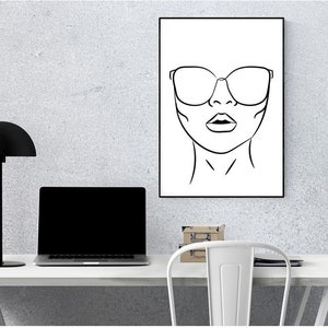 May include: Black and white line art print of a woman's face wearing sunglasses. The print is framed in a black frame and is hanging on a white wall.