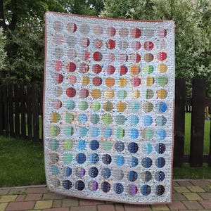 May include: A rectangular quilt featuring a grid of numerous circular fabric patches in a spectrum of colours including red, orange, yellow, green, blue, and purple. The quilt has a white background with small patterns and a thin red striped border.