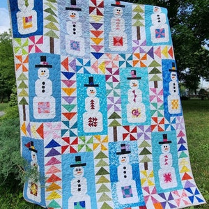 May include: A colorful quilt featuring a patchwork design of snowmen with various hats and accessories. The quilt is made with a variety of fabrics in shades of blue, green, red, yellow, and white. The quilt is displayed outdoors on a grassy lawn.