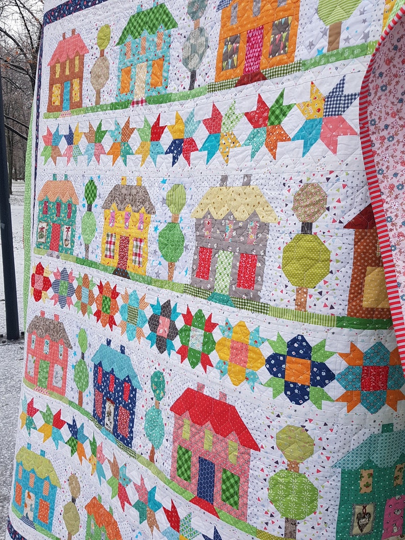 May include: A handmade quilt featuring rows of colorful houses and star patterns on a white background. The houses have various roof colors and door designs. The quilt's edge is green, and the backing has a floral pattern.