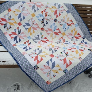 Op de afbeelding: Een kleurrijk quilt met een geometrisch patroon van witte, rode, blauwe, gele en roze driehoeken. De quilt heeft een blauw-witte ruitjesrand en ligt op een houten bank bedekt met sneeuw.