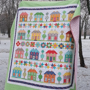 May include: A colorful quilt featuring rows of houses, trees, and star patterns. The quilt has a white background with houses in various colors like blue, orange, and green. The border is green with a floral pattern on the reverse side.