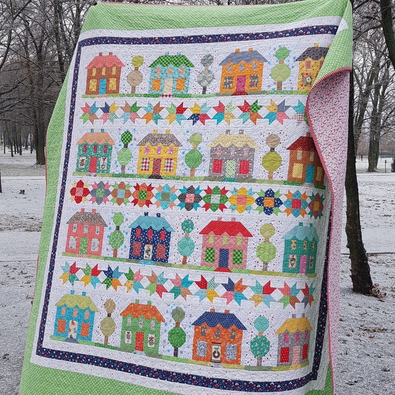 May include: A colorful quilt featuring rows of houses, trees, and star patterns. The quilt has a white background with houses in various colors like blue, orange, and green. The border is green with a floral pattern on the reverse side.
