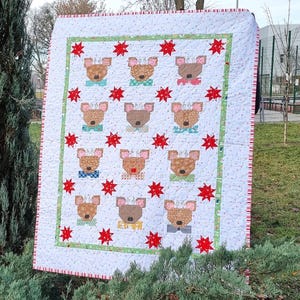 Può includere: Un quilt bianco con un bordo rosso e verde. Il quilt presenta un motivo ripetitivo di volti di renne marroni con nasi rossi e corna bianche. Le renne sono circondate da stelle rosse.