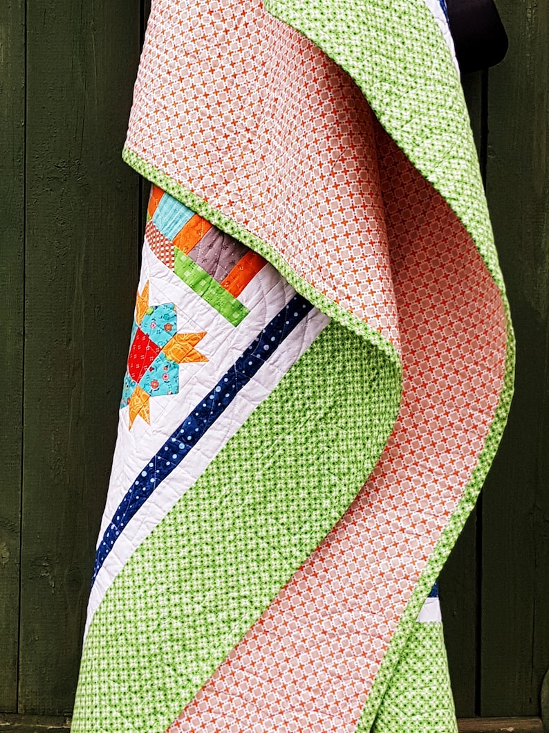 May include: A colorful quilt with a starburst design, featuring orange, green, blue, and white patterns. The quilt has a patchwork design with a variety of geometric shapes and textures. The quilt is folded over, showcasing the different patterns and colors.