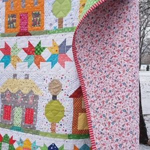 May include: A colorful patchwork quilt featuring houses, trees, and star patterns. The quilt showcases orange, gray, and brown houses with patterned roofs and doors. The reverse side has a floral pattern with a red and white striped border.