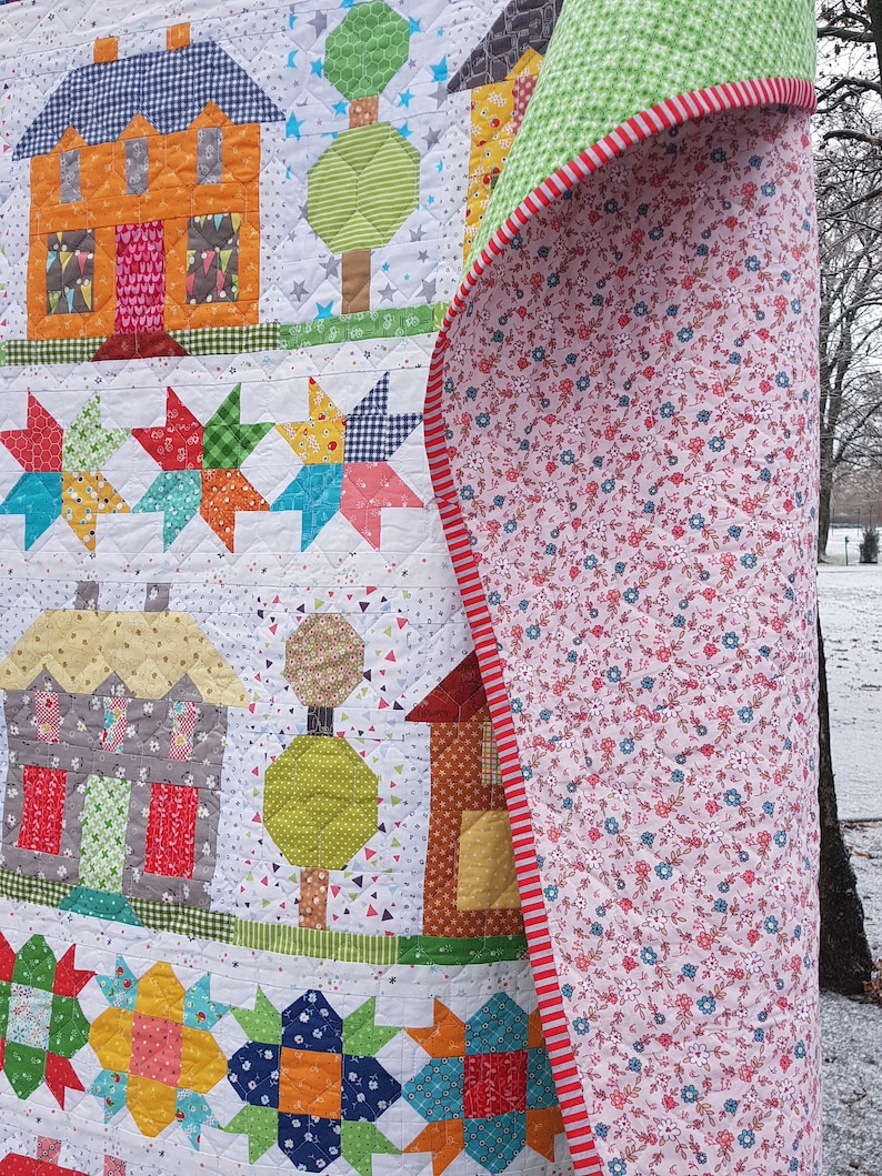 May include: A colorful patchwork quilt featuring houses, trees, and star patterns. The quilt showcases orange, gray, and brown houses with patterned roofs and doors. The reverse side has a floral pattern with a red and white striped border.