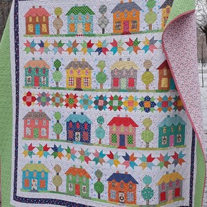 May include: A handmade quilt featuring a repeating pattern of colorful houses and trees on a white background. The quilt has a patchwork design with star and floral accents. The edges are finished with a green border and a floral patterned backing.